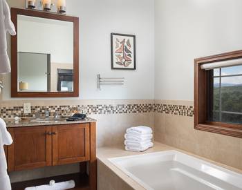 Wood cabinet holding a marble top bathroom sink, soaking tub with a window above with a view of the Mountains